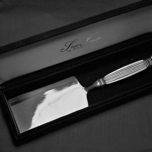 May include: A silver cake server in a black velvet-lined box. The server has a wide, rectangular blade and a decorative handle with vertical grooves and ornate detailing. The box has the text "Silversmiths by Gorham".