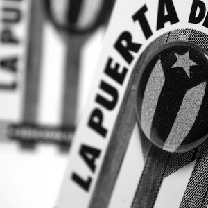 May include: A black and white button with the text "La Puerta De La Bandera" and a stylized image of the Puerto Rican flag.