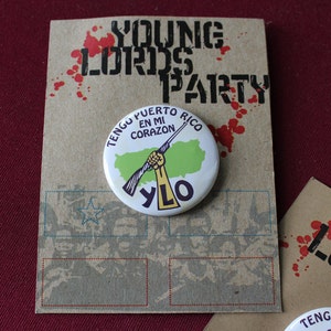 May include: A brown paper card with the text "Young Lords Party" in black lettering. The card has red splatter paint and a white button with the text "Tengo Puerto Rico En Mi Corazon" and a green outline of Puerto Rico with a gun and fist. The button also has the text "YLO" in black lettering.
