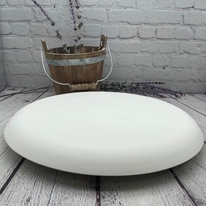 May include: A large, white, round ceramic platter with a slightly raised edge. The platter is sitting on a light wood surface.