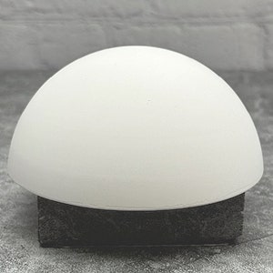 May include: A white ceramic dome sits on a black square base. The dome is smooth and rounded, with a slightly textured surface.