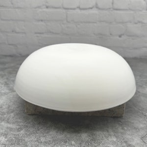 May include: A white ceramic bowl with a smooth, rounded surface. The bowl is sitting on a rough, textured surface.