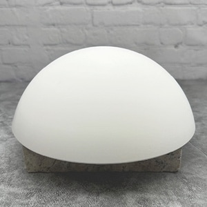 May include: A white, semi-spherical object, possibly a lampshade or decorative element, rests on a rectangular, textured, gray stone base. The smooth, curved surface of the object contrasts with the rough texture of the base.