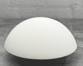 Könnte beinhalten: Eine weiße, kuppelförmige Lampe mit einer glatten, abgerundeten Oberfläche. Die Lampe steht auf einer grauen Oberfläche.