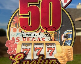 May include: A red and gold 50th birthday cake topper with a Las Vegas theme. The topper features a slot machine with the number 777, playing cards, and a sign that says "Welcome to Fabulous Las Vegas". The name "Evelyn" is written in gold letters on the bottom of the topper.