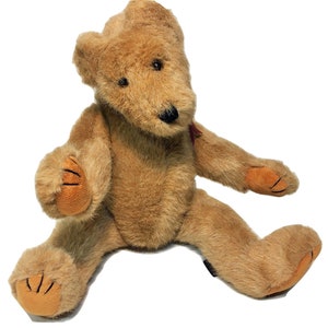 Puede incluir: Un oso de peluche marrón claro sentado, con patas de color canela. El oso tiene una nariz negra y ojos pequeños negros. Una pequeña cinta roja atada alrededor del cuello. Un juguete infantil clásico.