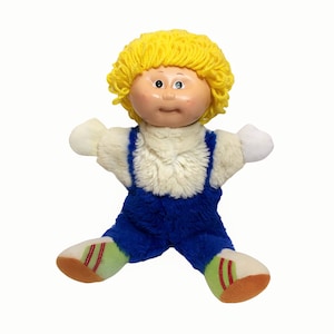 May include: A plush doll with a light skin tone, yellow yarn hair, and blue overalls. The doll has a white fluffy shirt and white hands. The doll's feet are white with green and red accents. The doll is a vintage toy.