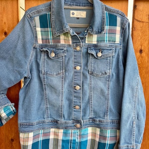 May include: A blue denim jacket featuring a plaid pattern in teal, white, and red on the upper chest panels and the lower hem. The jacket has a button-front closure, two chest pockets with button flaps, and frayed cuffs revealing the plaid fabric.