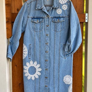 May include: A long-length light blue denim jacket featuring a button-down front, two chest pockets, and a collar. The jacket is decorated with various white circular crochet lace doilies of different sizes applied to the shoulders and lower body.
