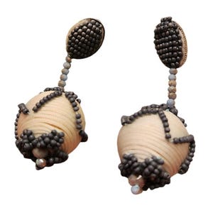 May include: A pair of dangle earrings with a cream colored ball wrapped in a beaded design and a small oval shaped bead at the top. The beads are dark gray and light blue.