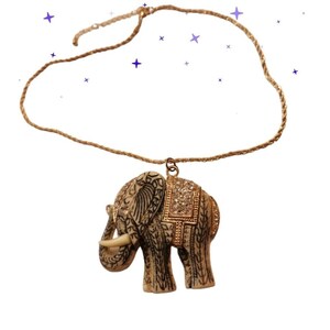 May include: A necklace featuring a pendant shaped like an elephant with a cream-colored body, black etched patterns, and a gold-toned saddle adorned with small clear crystals. The pendant hangs from a thin, gold-colored twisted cord chain.