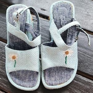 May include: A pair of light blue woven sandals with a daisy embroidery on the front. The sandals have a strap that buckles around the ankle. The soles are a darker blue color.