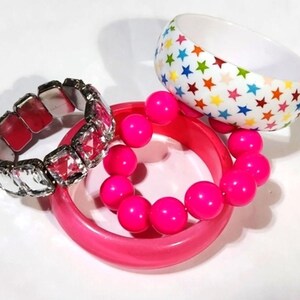 May include: A collection of bracelets in various styles. One bracelet features large, clear, faceted stones with a silver-tone metal setting. Another is a wide, white bangle with colorful star patterns. A third bracelet is a pink bangle with pink beads.