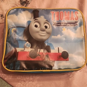 May include: A blue and red Thomas the Tank Engine lunch bag with a yellow zipper. The bag features a cartoon image of Thomas with a blue and white engine and a red caboose. The text "THOMAS THE TANK ENGINE" is printed in red and white on the bag.
