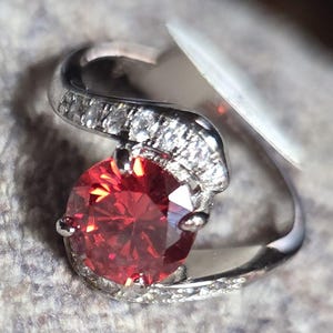 May include: A silver ring with a large, oval-cut, red gemstone as the focal point. The band curves around the stone, with smaller clear stones set along the top curve. The ring is set against a neutral background.