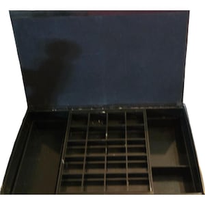 May include: A black jewelry storage box with an open lid featuring a dark blue interior lining. The base contains a central removable tray with multiple small square compartments, flanked by two larger open storage sections on either side.