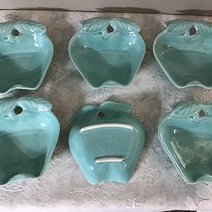 May include: Set of six teal green ceramic apple-shaped bowls. Each bowl has a leaf design and a small hole in the top. One bowl has the text "Homer Laughlin U.S.A." on the bottom.