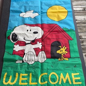 May include: A rectangular fabric garden flag featuring a blue sky with a yellow sun and white cloud, a red doghouse, and a green lawn. The white dog Snoopy stands in front of the doghouse with a yellow bird, and the word WELCOME is printed in yellow.