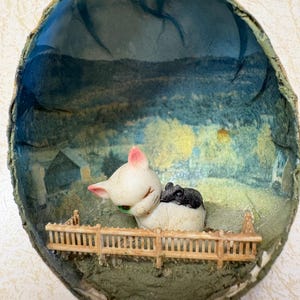 May include: A diorama featuring a white cat with pink ears and a black and white mouse, set against a painted landscape. The scene includes a miniature wooden fence and a backdrop of a field and a house.