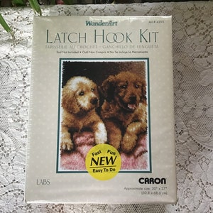 May include: Latch hook kit featuring two golden retriever puppies on a pink blanket. The kit includes yarn, canvas, and instructions. The finished project will be approximately 20 inches by 27 inches.