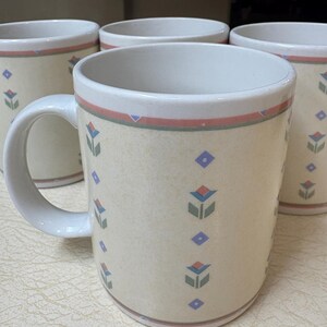 May include: Set of four cream-colored ceramic mugs. Each mug features a floral and geometric pattern in blue, red, and green. The mugs have a white interior and a handle for easy gripping. The mugs are suitable for hot or cold beverages.