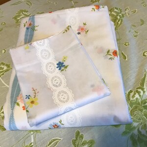May include: Two white pillowcases with a floral pattern and lace trim. The pillowcases are folded and placed on a green floral patterned fabric.