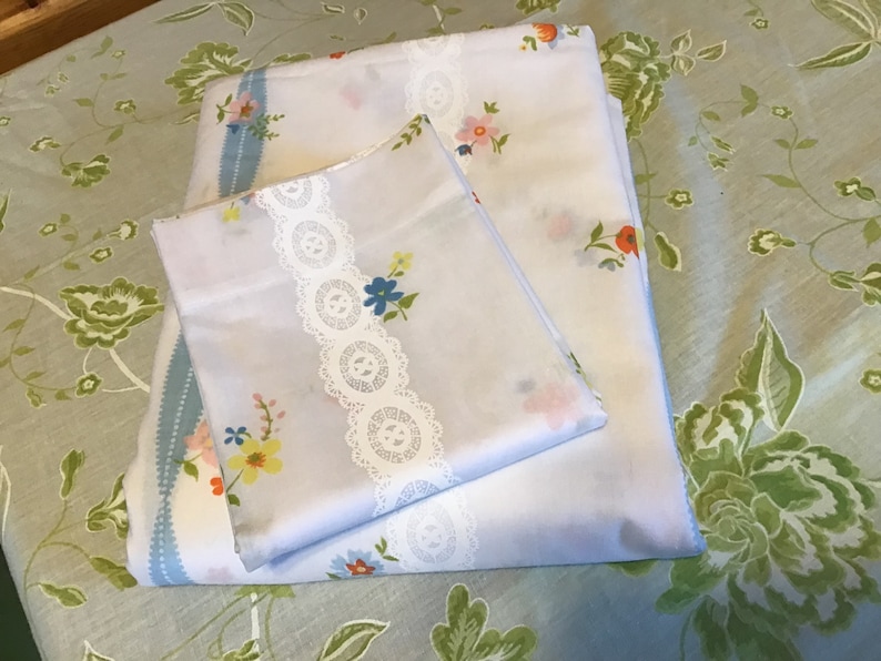 May include: Two white pillowcases with a floral pattern and lace trim. The pillowcases are folded and placed on a green floral patterned fabric.