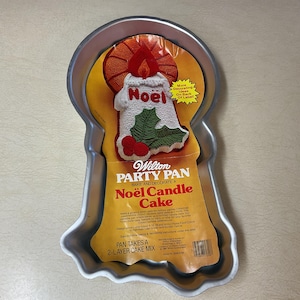 May include: A silver-colored Wilton Party Pan in the shape of a candle. The pan has a yellow and orange label with the words "Noël Candle Cake" and a decorative candle design. The pan takes a 2-layer cake mix.