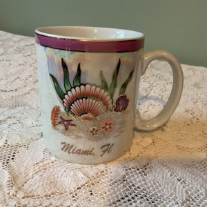 May include: White ceramic mug with a pink and gold rim. The mug features a seashell design with green seaweed and a starfish on a sandy beach. The text "Miami, Fl" is printed on the bottom of the mug.