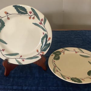 May include: Two ceramic plates with a cream-colored background. One is round and propped on a wooden stand, the other is oval. Both plates feature a green leaf and red berry design around the edges. The plates are suitable for serving food.