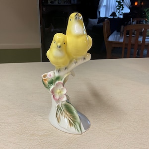 May include: A ceramic figurine of two yellow birds perched on a branch with pink flowers and green leaves. The birds are facing different directions, and the figurine has a glossy finish. The base is white and gray.