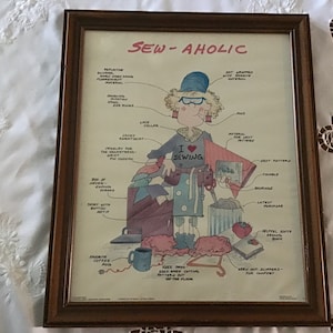 May include: A humorous illustration of a woman who loves to sew. The woman is wearing a red shirt with the words "I love sewing" on it. She is surrounded by sewing supplies, including fabric, thread, scissors, and a sewing machine. The caption reads "Sew-aholic".