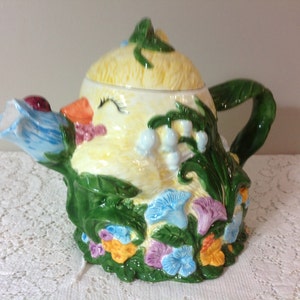 May include: A yellow ceramic teapot shaped like a chick with a blue flower spout and green leaves. The teapot is decorated with flowers and a ladybug.