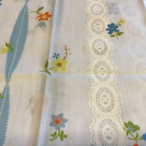 May include: White fabric with a blue and white pattern and a white lace trim. The fabric has a floral design with pink, yellow, blue, and orange flowers.