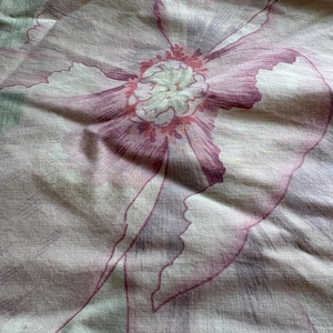 May include: A textile fabric featuring a large-scale floral pattern in shades of muted mauve, dusty pink, and sage green. The design displays a central flower with intricate petal details and a textured appearance, printed on a light-colored base.