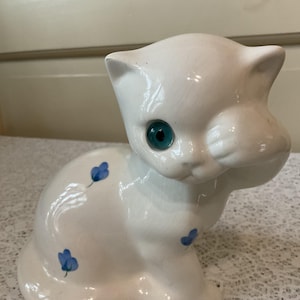 May include: White ceramic cat figurine with blue eyes and blue floral accents. The cat is sitting with its head tilted and one paw raised.