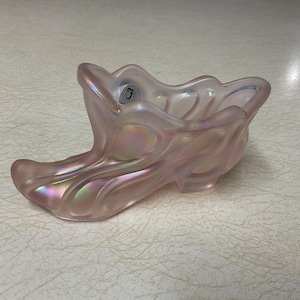 May include: A decorative, iridescent pink glass slipper-shaped dish. The dish has a wavy rim and a textured, swirling design. The glass reflects a rainbow of colors. A small, circular sticker is visible on the rim.