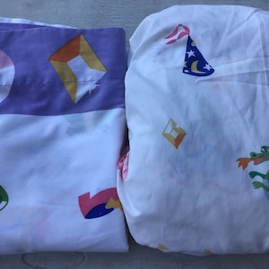 May include: Two white twin-sized bed sheets with colorful cartoon designs. One sheet features a pink star, a yellow diamond, and a green eye. The other sheet features a blue and white hat, a yellow rectangle, and a green dragon.