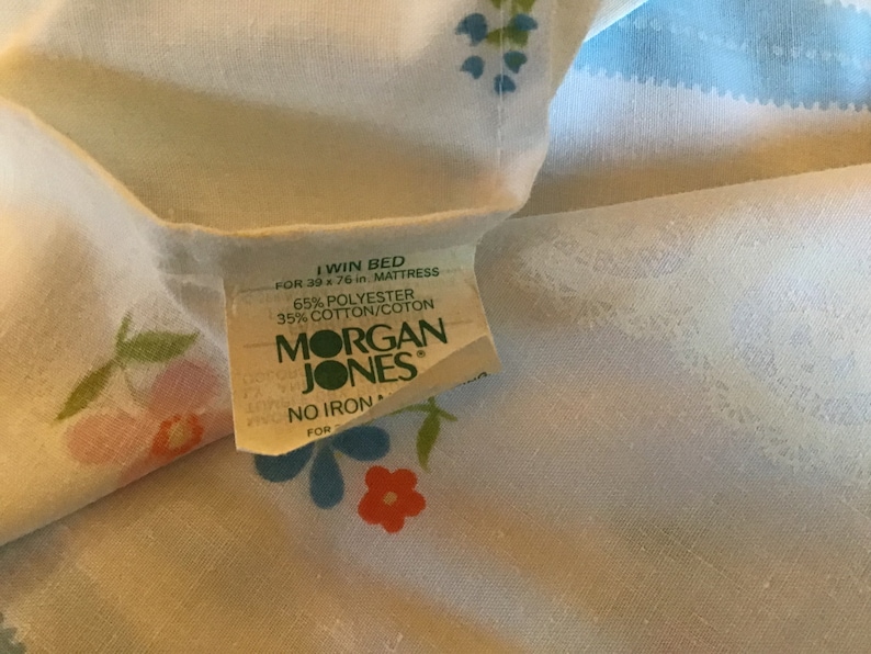 May include: A white twin bed sheet with a floral pattern in shades of pink, blue, and green. The sheet has a label that reads "I Win Bed for 39 x 76 in. Mattress 65% Polyester 35% Cotton/Coton Morgan Jones No Iron For"