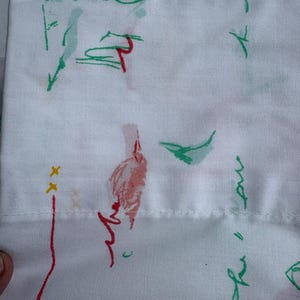 May include: A white fabric featuring a scattered pattern of hand-drawn style sketches in red, green, and yellow. The design includes abstract shapes, thin lines, and small cross marks, with a horizontal stitched seam running across the lower section.