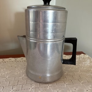 May include: A vintage aluminum coffee percolator with a black handle and lid knob. The coffee pot is cylindrical with a spout and a lid. The percolator is sitting on a white lace tablecloth.