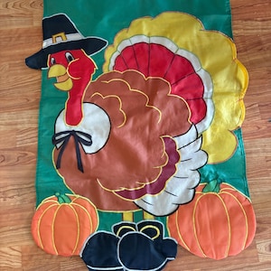 May include: A rectangular green garden flag featuring a central turkey illustration wearing a black pilgrim hat and a white collar with a black bow. The turkey has brown, red, and yellow feathers, flanked by two orange pumpkins at the base.
