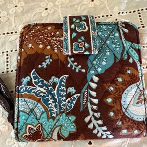 May include: A square quilted wallet featuring a dark brown base with a vibrant floral and paisley pattern in shades of teal, light blue, and cream. The wallet includes a matching fabric strap closure and a small side zipper pull with visible text.