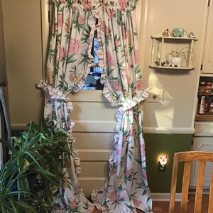May include: A pair of white floral curtains with ruffled edges hang in a doorway. The curtains are tied back with a black and white checkered ribbon. A green plant is in the foreground.