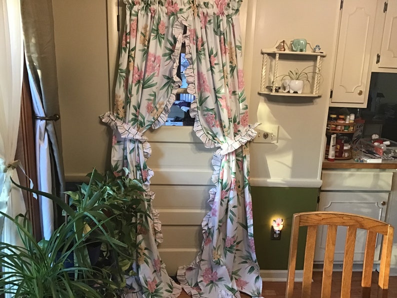 May include: A pair of white floral curtains with ruffled edges hang in a doorway. The curtains are tied back with a black and white checkered ribbon. A green plant is in the foreground.