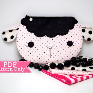 May include: A pink and black polka dot lamb-shaped zippered pouch with a black felt face and a white pearl zipper pull.  The pouch is sitting on a white surface with black beads and pink and white fabric.