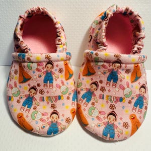May include: A pair of pink baby shoes with a colorful pattern of dolls, bunnies, and the words "Hi Friends".