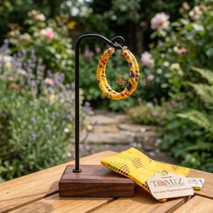 Peut inclure: Boucles d'oreilles créoles en tissu jaune avec un motif coloré, présentées sur un support noir avec une base en bois. Un tissu jaune assorti est posé à côté. L'arrière-plan est un jardin avec des fleurs et de la verdure.