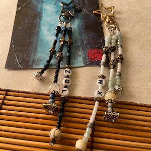 Puede incluir: Dos llaveros de cuentas con dijes en forma de estrella. Un llavero es negro con cuentas azules y un dije con la letra "Z". El otro llavero es blanco con cuentas azul claro y un dije con la letra "W".