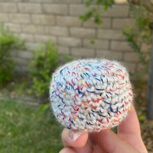 May include: A small, spherical object crafted from white yarn with a multi-colored speckled pattern. The item features a dense, textured crochet stitch throughout its rounded surface, creating a uniform and compact appearance in a ball shape.